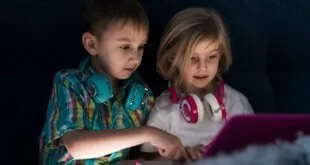 Duas crianças com fones de ouvido no pescoço olham a tela iluminada de um tablet