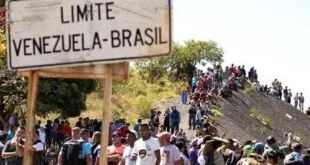 Placa de estrada com os dizeres 'Limite Venezuela-Brasil' e fila de pessoas caminhando