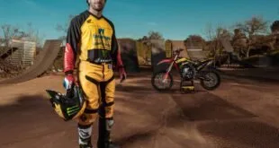 Piloto de motocross com uniforme amarelo segura o capacete diante da moto e de rampas
