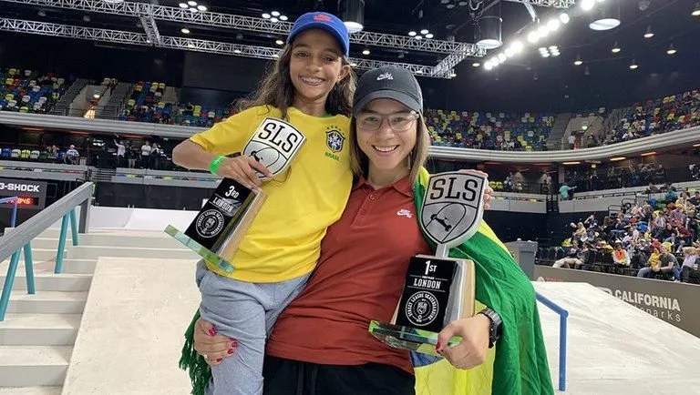 Duas skatistas com medalhas e troféus enroladas em bandeira do Brasil dentro de arena