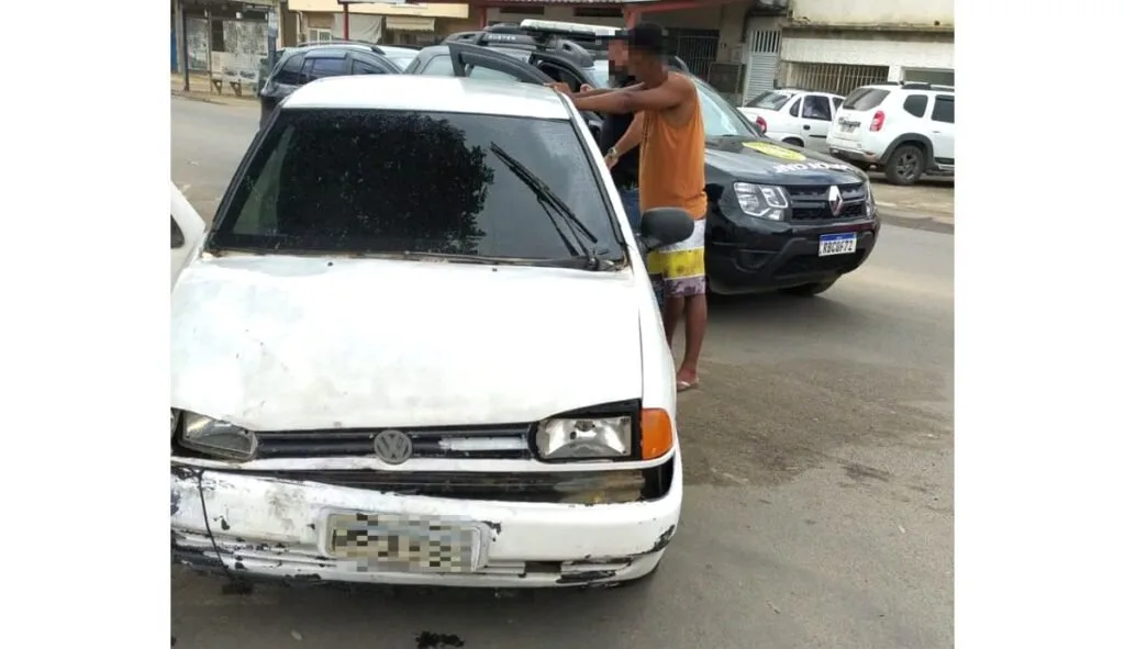 Homem ao lado de carro branco danificado com viaturas policiais ao fundo na rua