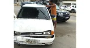 Homem ao lado de carro branco danificado com viaturas policiais ao fundo na rua