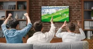 Casal de braços erguidos comemora diante de televisão que exibe partida de futebol