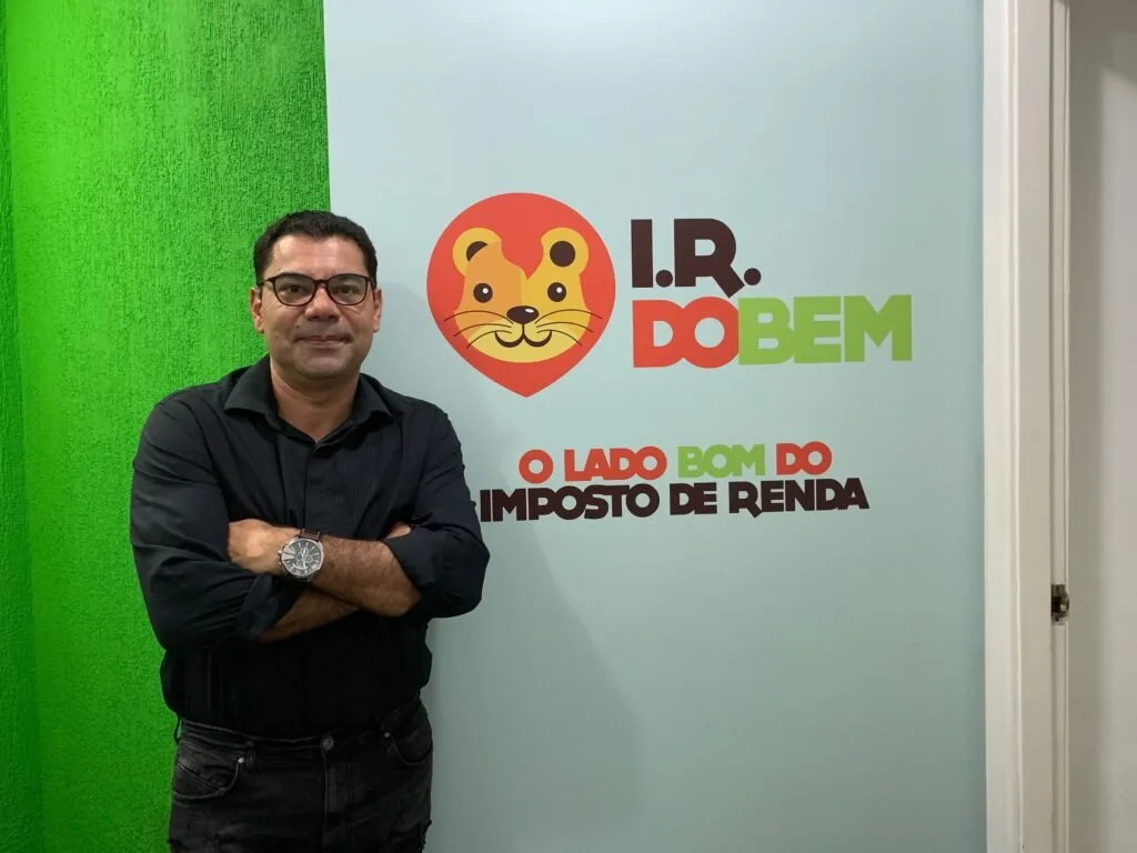 Homem de braços cruzados diante de parede com o logotipo 'I.R. do Bem'