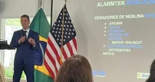 Homem de terno fala ao microfone entre bandeiras do Brasil e dos EUA diante de slide da Alarmtek