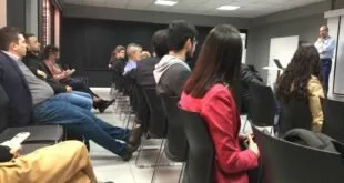 Plateia sentada em cadeiras acompanha palestrante em sala de reunião