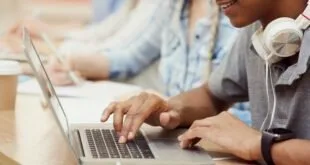 Mãos digitando em notebook em sala com outras pessoas ao lado