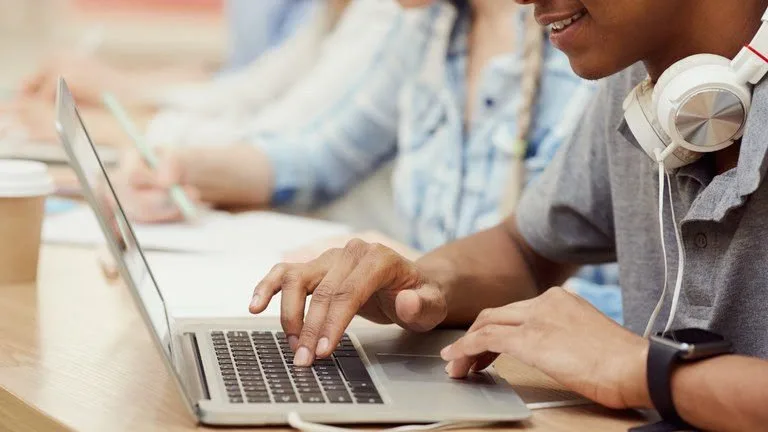 Mãos digitando em notebook em sala com outras pessoas ao lado