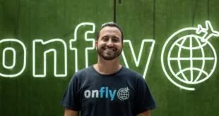 Homem sorrindo diante de letreiro em neon com o nome 'onfly' em parede verde