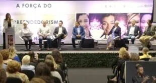 Pessoas sentadas em palco de evento diante de plateia, com telões ao fundo