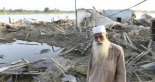Homem idoso de barba branca diante de casas destruídas e área alagada