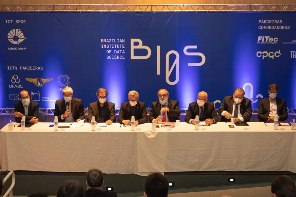 Homens de terno e máscara sentados à mesa diante de painel do Brazilian Institute of Data Science