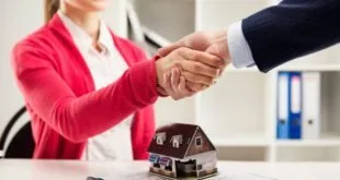 Aperto de mãos sobre mesa com miniatura de casa e documentos ao lado