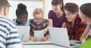 Estudantes sorrindo em volta de mesa com notebooks e tablet ao lado de professora
