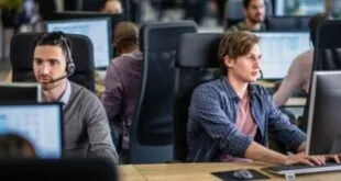 Jovem sentado diante de computadores em escritório aberto com colegas ao fundo