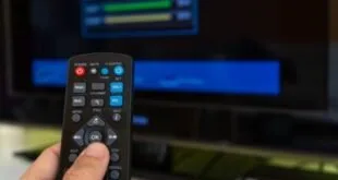 Mão segurando controle remoto diante de televisão e aparelho ligados no escuro