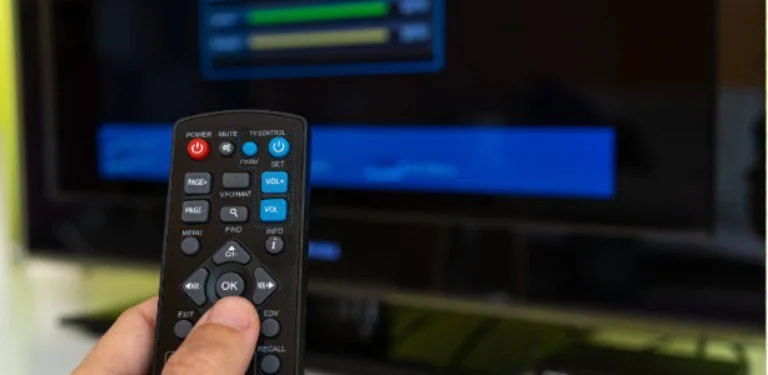 Mão segurando controle remoto diante de televisão e aparelho ligados no escuro