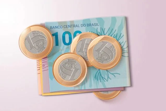 Moedas de um real sobre cédula de cem reais em fundo rosa