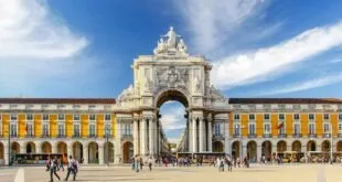 Arco monumental de pedra entre prédios amarelos com bandeiras de Portugal e pessoas na praça
