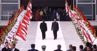 Dois homens subindo rampa com tapete branco entre autoridades e arranjos de flores