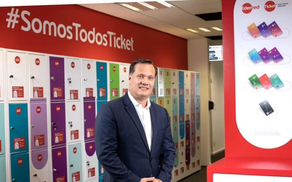 Homem de terno em pé diante de parede vermelha com o letreiro 'Ticket' e vitrine de produtos