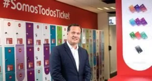 Homem de terno em pé diante de parede vermelha com o letreiro 'Ticket' e vitrine de produtos