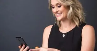Mulher sentada em cadeira de madeira sorrindo enquanto usa o celular diante de fundo escuro