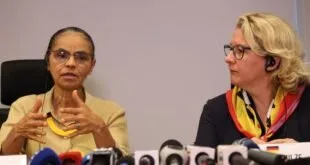 Duas mulheres sentadas diante de vários microfones de rádio e TV em entrevista coletiva