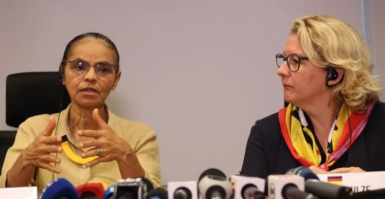 Duas mulheres sentadas diante de vários microfones de rádio e TV em entrevista coletiva