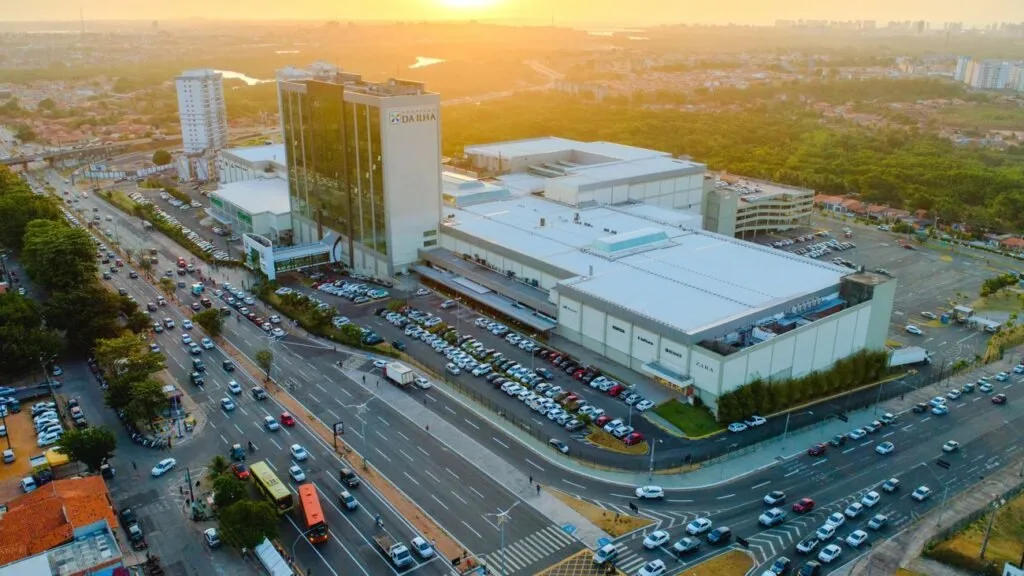 Vista aérea de complexo de shopping center com estacionamento cheio e avenida movimentada ao pôr do sol