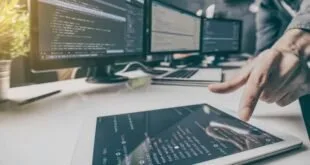 Pessoa diante de monitores com códigos e tablet com gráficos sobre a mesa