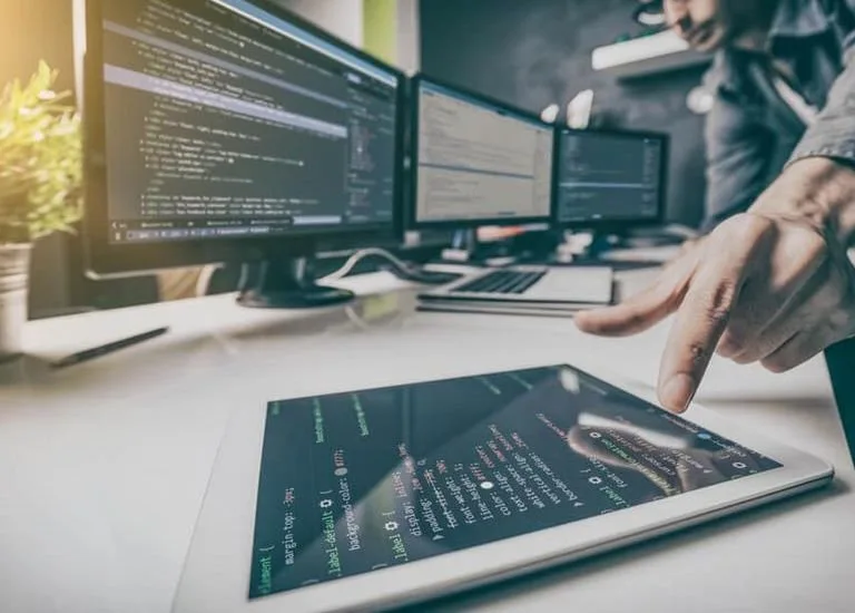 Pessoa diante de monitores com códigos e tablet com gráficos sobre a mesa
