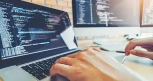 Mãos digitando em notebook com linhas de código na tela e monitor ao fundo