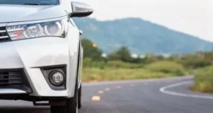 Frente de carro branco parado à beira de estrada asfaltada com morros ao fundo