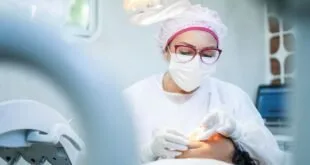 Dentista de máscara, touca e óculos atende paciente deitado em consultório odontológico