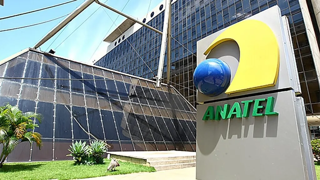 Totem com o logotipo e o nome 'ANATEL' em frente a prédio de fachada de vidro espelhado