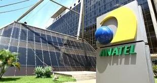 Totem com o logotipo e o nome 'ANATEL' em frente a prédio de fachada de vidro espelhado