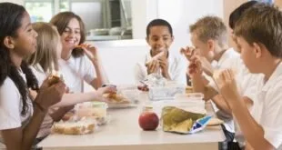 Crianças sentadas à mesa da escola comendo lanches, com frutas e potes plásticos à frente