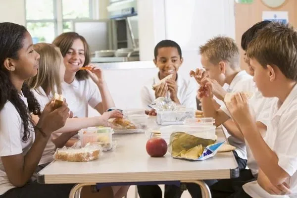Crianças sentadas à mesa da escola comendo lanches, com frutas e potes plásticos à frente