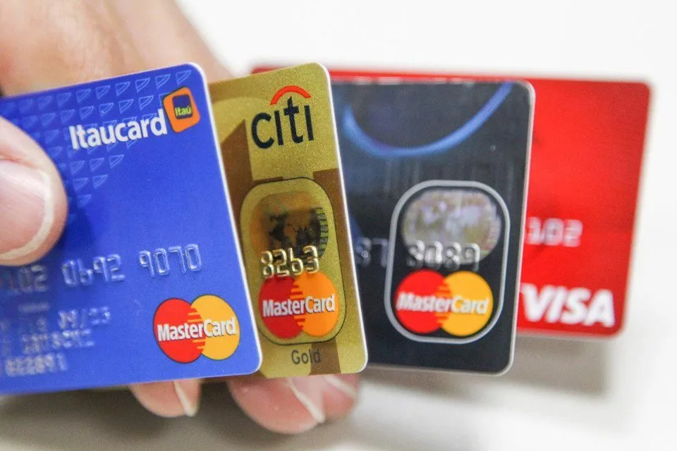 Mão segurando três cartões de crédito em leque, com bandeiras e números em relevo