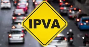 Placa amarela de trânsito com a sigla IPVA diante de avenida com carros desfocados