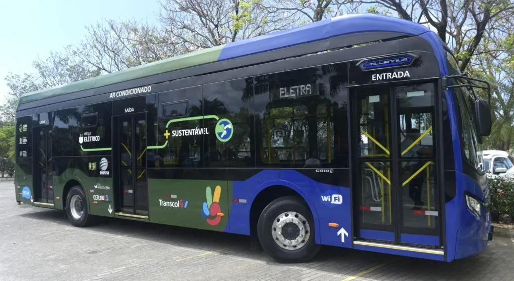 Ônibus elétrico azul e verde com a marca Transcol na lateral, estacionado sob árvores
