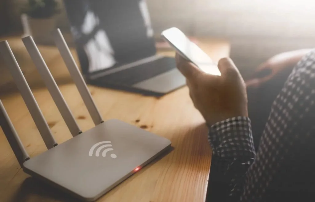 Roteador de Wi-Fi branco com quatro antenas sobre mesa de madeira e pessoa segurando celular ao lado