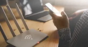 Roteador de Wi-Fi branco com quatro antenas sobre mesa de madeira e pessoa segurando celular ao lado