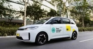 Carro branco de aplicativo com adesivo 'Zero emissão de CO2' e número 99 trafegando em avenida arborizada