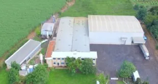 Vista aérea de galpões industriais cercados por plantação verde, com carreta no pátio