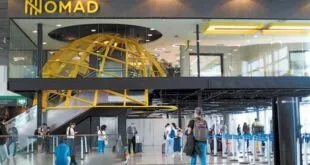 Interior de terminal com letreiro amarelo iluminado e pessoas caminhando sob estrutura de vidro