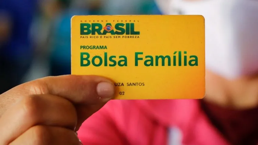 Mão segurando cartão amarelo do programa Bolsa Família com o brasão do Brasil