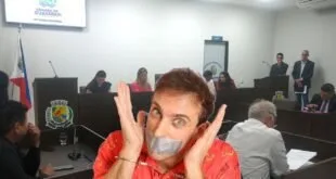 Homem com fita adesiva sobre a boca e mãos erguidas ao lado do rosto em sala de sessões