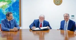 Homem assina documento em mesa entre dois outros homens de terno, em sala oficial com brasão na parede